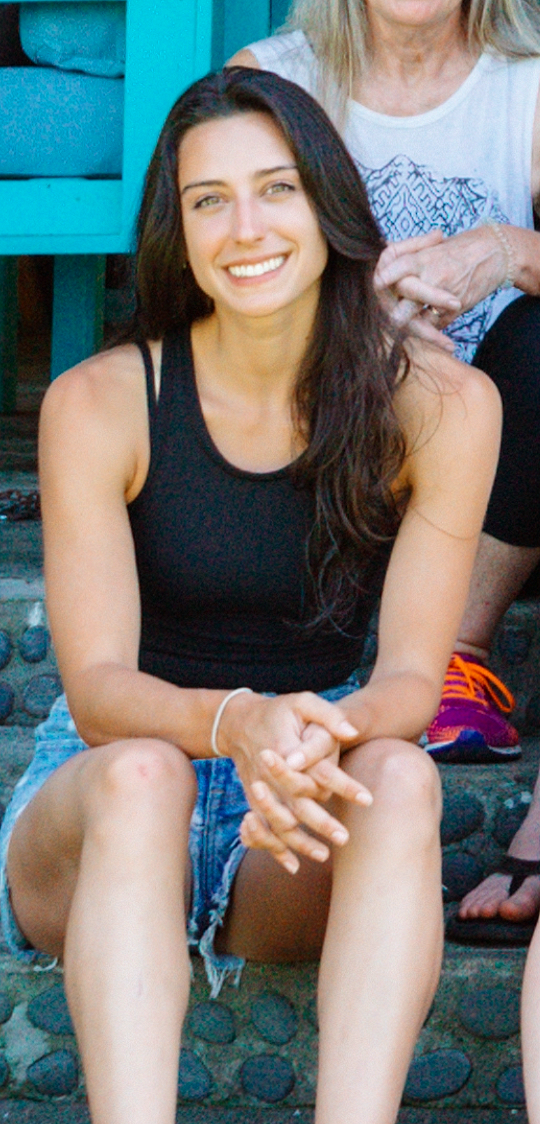 Young woman with long dark hair smiling, sitting outdoors on a stone surface, wearing a black tank top and denim shorts, with another person partially behind her.