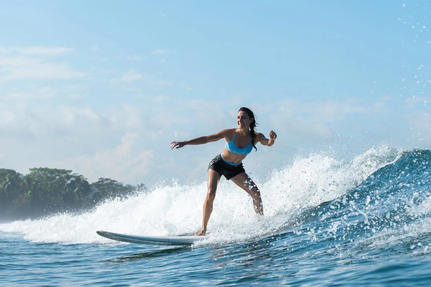 A woman surfing on a wave in the ocean, smiling and wearing a blue sports bra and black shorts.