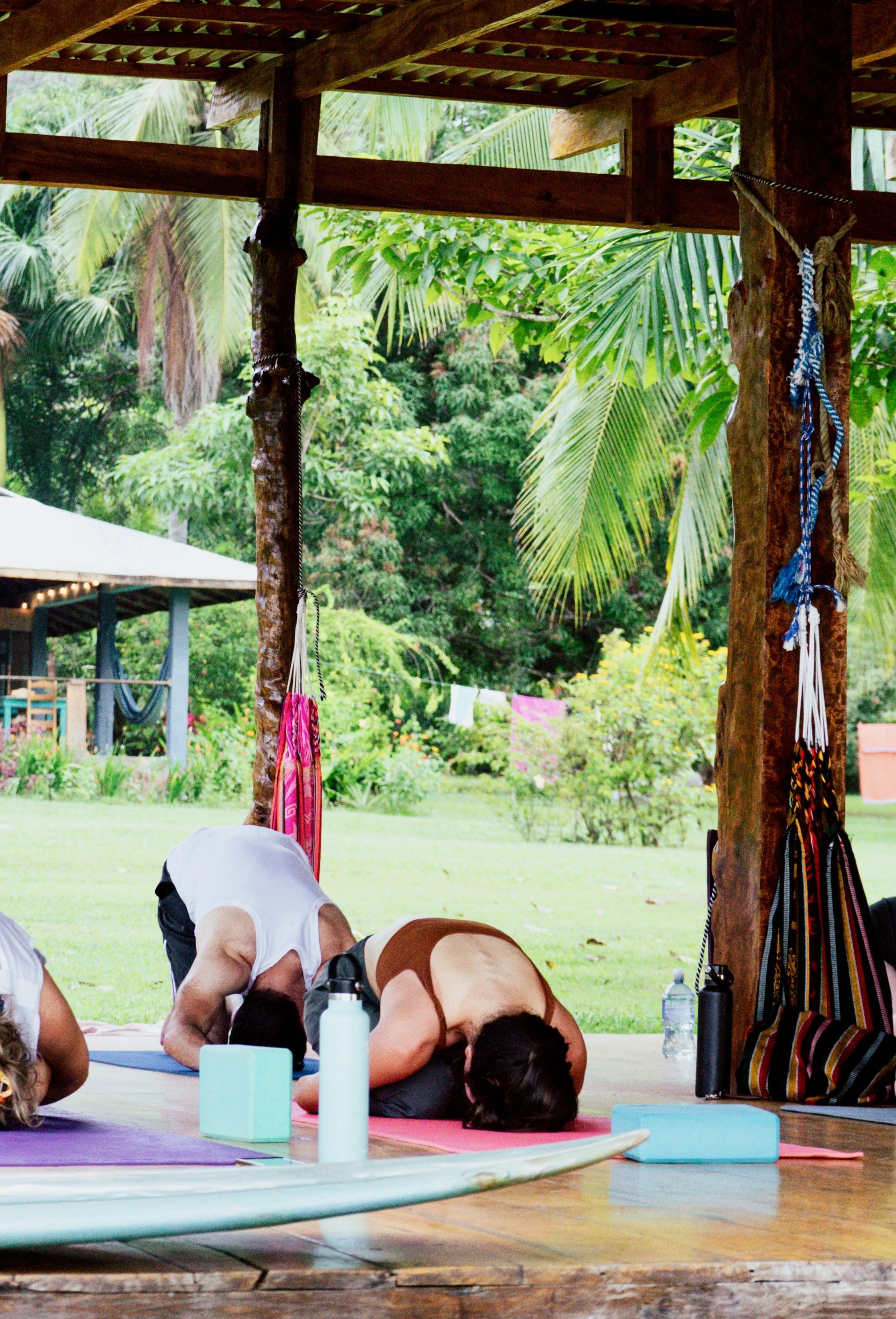 People doing yoga or stretching exercises under a wooden shelter in a lush green outdoor setting.