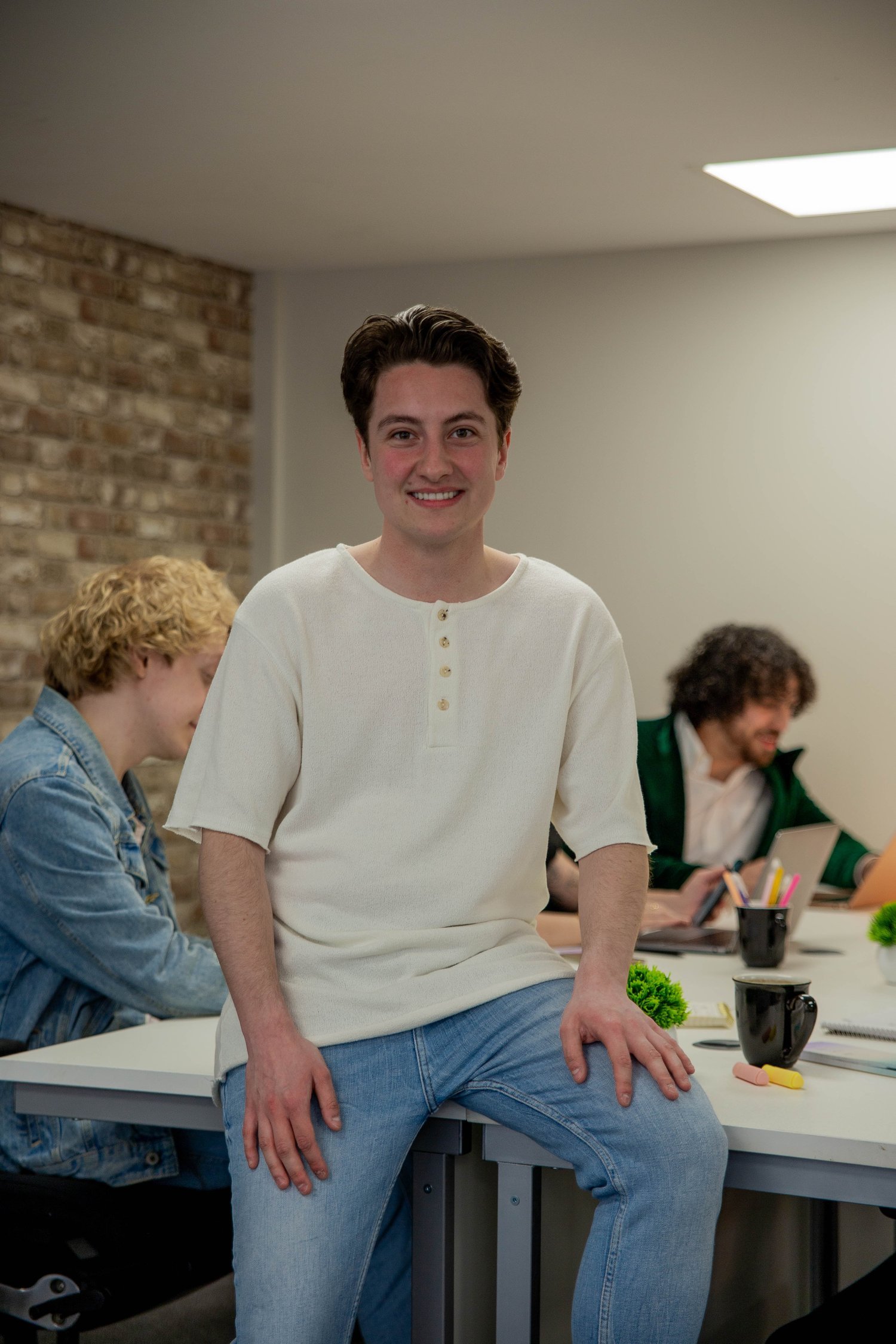 Image of Tate Smith, a dark haired, white male seated towards the camera, perched on a desk, smiling towards the camera.