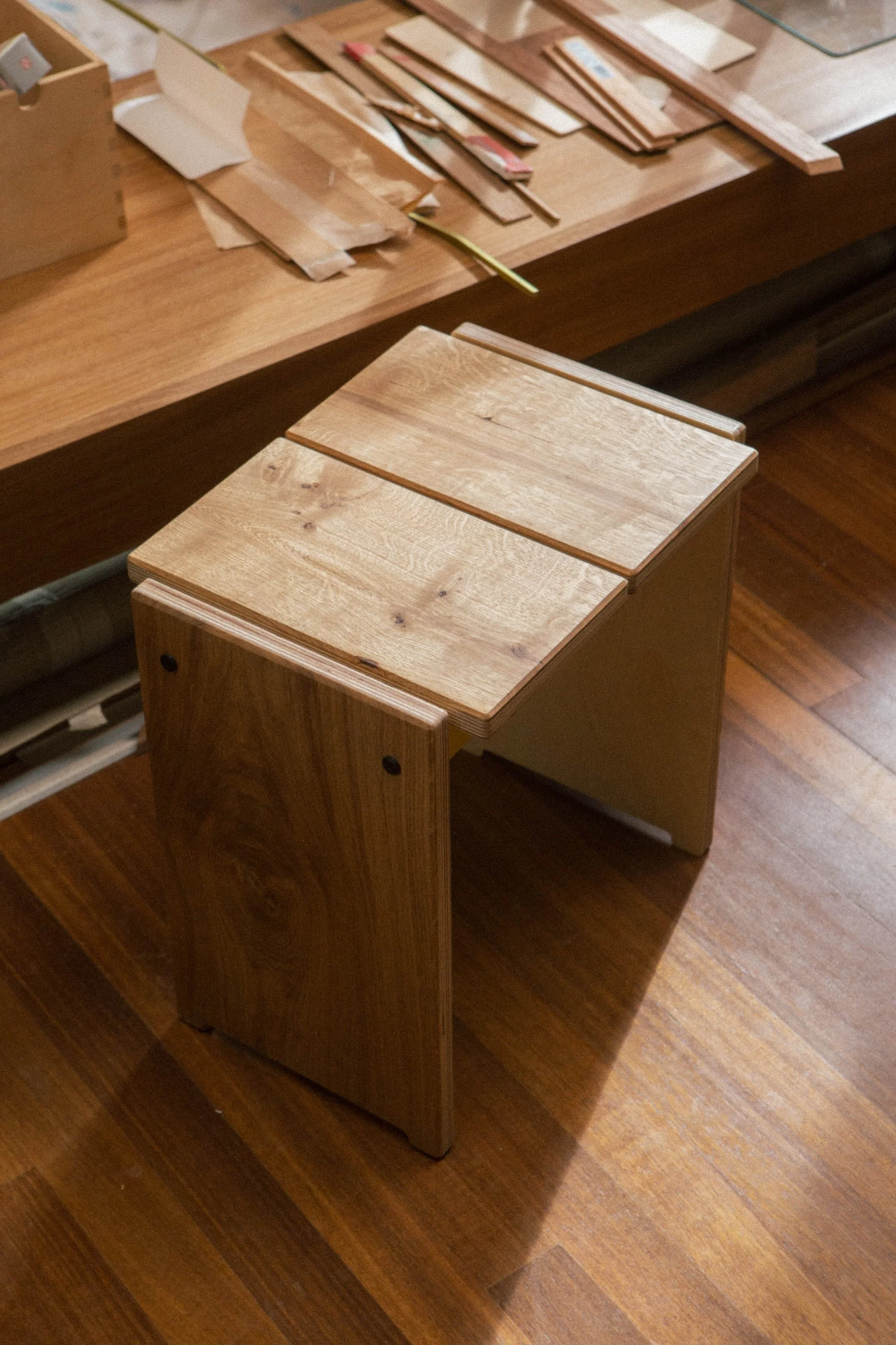 A wooden stool with a unique design, featuring two tops, placed on a hardwood floor near a wooden table with various pieces of wood and tools on it.