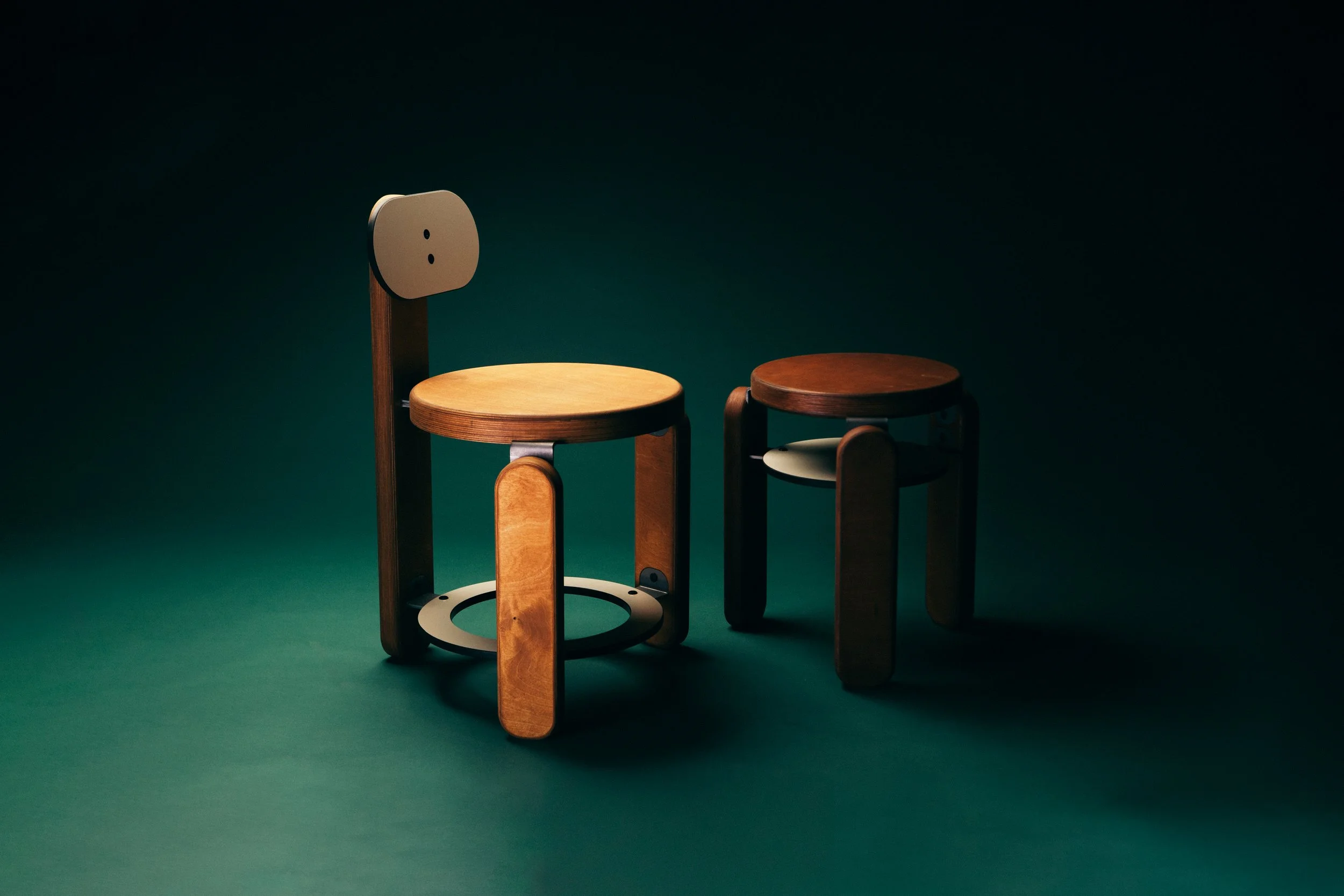 Two wooden chairs with rounded legs and circular seats, one with a backrest resembling a face, against a dark green background.