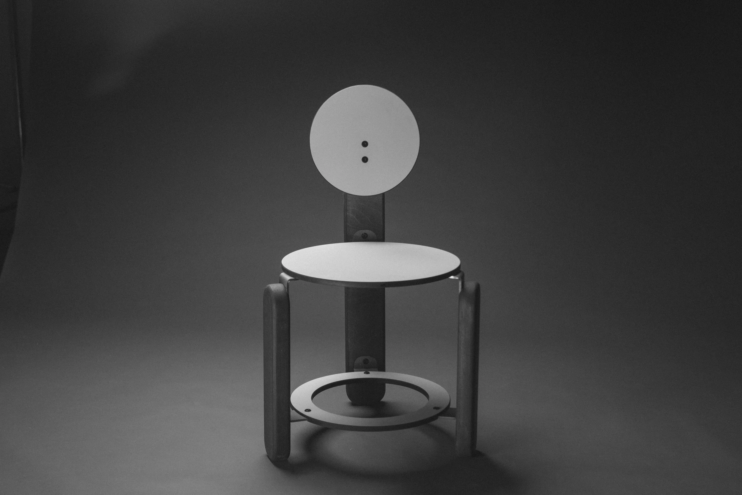 A modern chair with wooden side supports, a round seat, and a backrest that is a flat circle with two small holes in the center, set against a dark background.