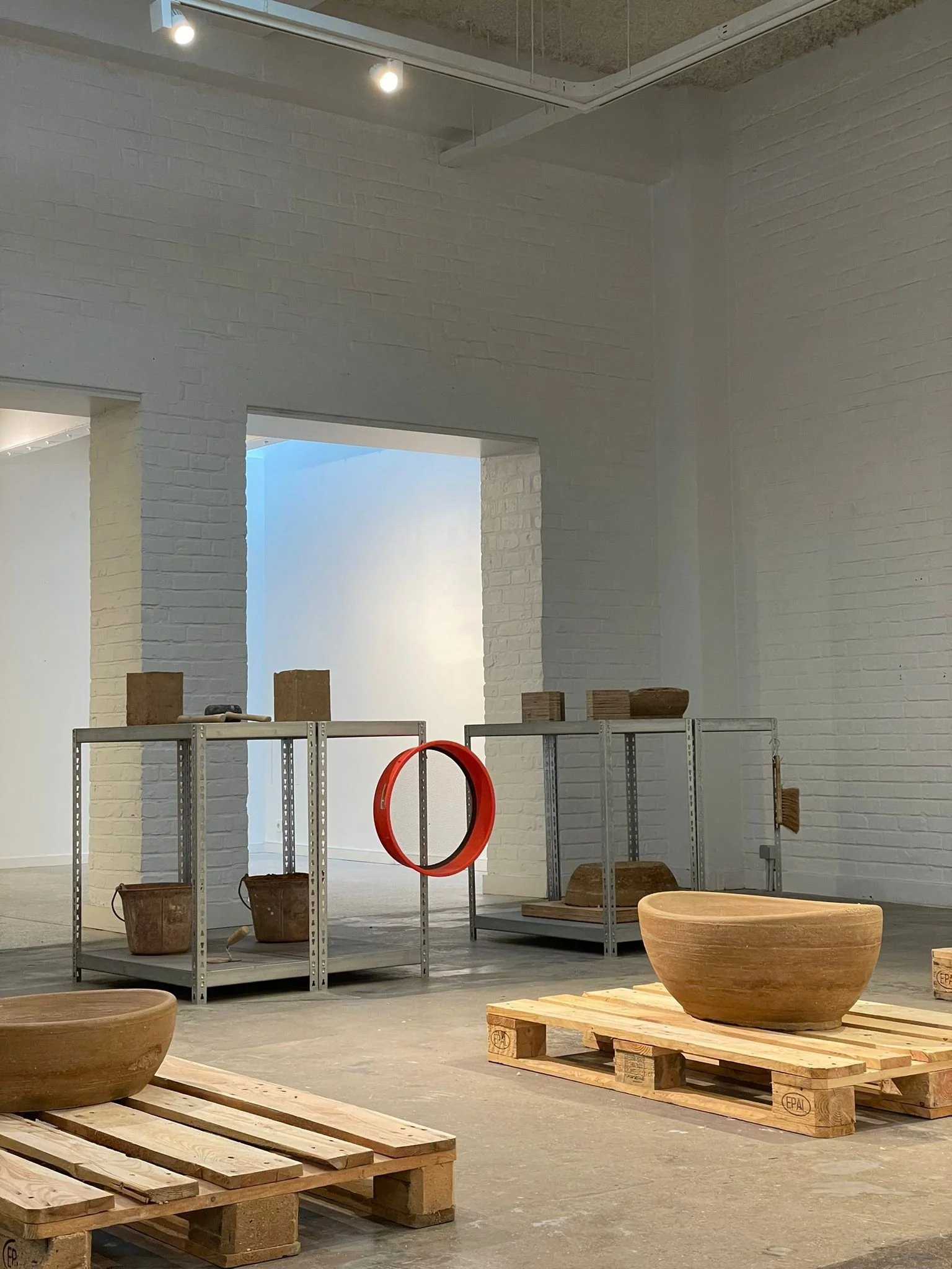 Interior of an art gallery or studio with white brick walls and industrial ceiling, displaying various wooden bowls and blocks on pallets and metal shelves.