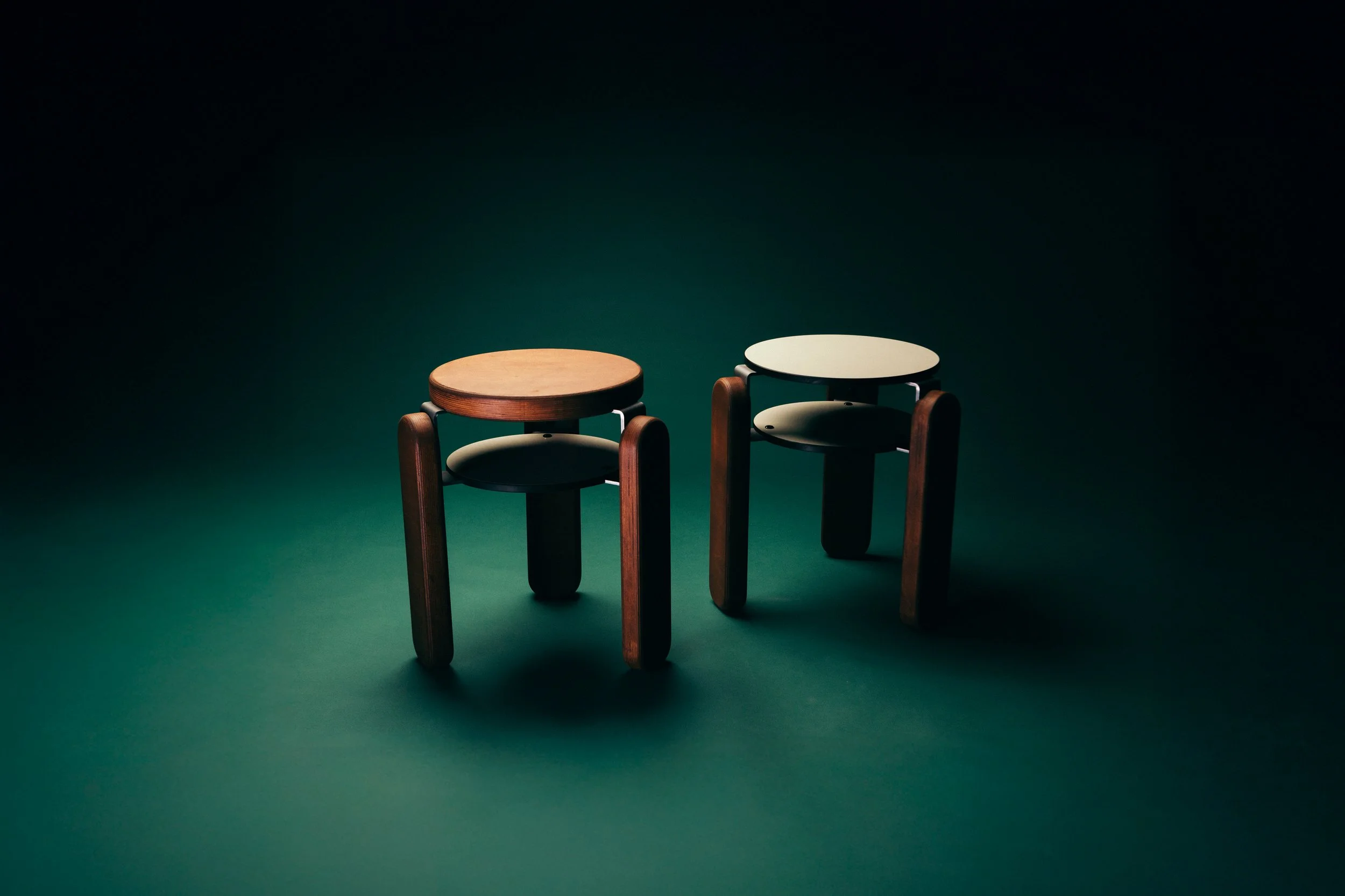 Two small wooden stools with round tops, one in natural wood color and the other painted white, are placed on a dark green surface with a dark background.
