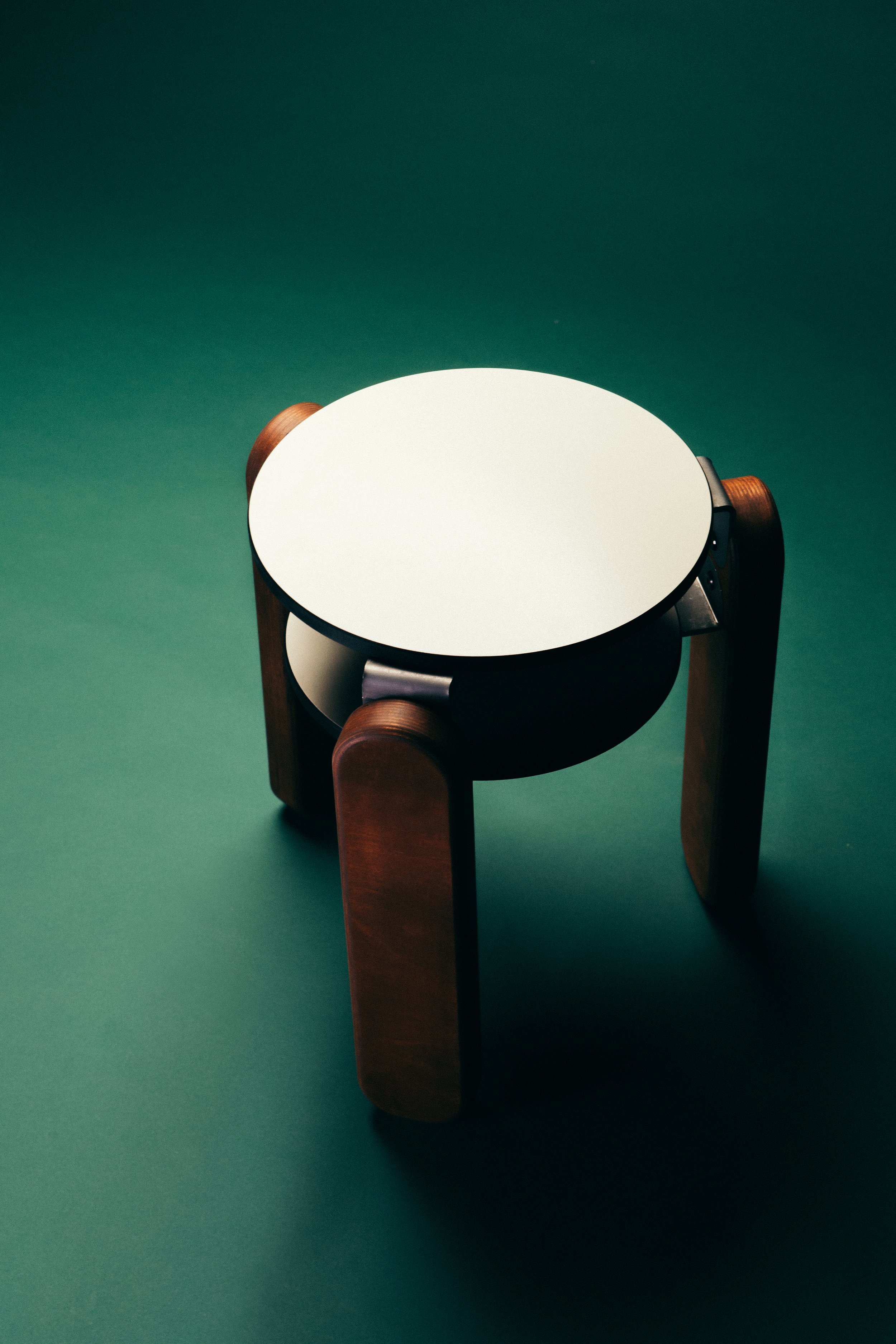 A small round white table with wooden legs placed on a green surface. The lighting creates shadows on the table and floor.