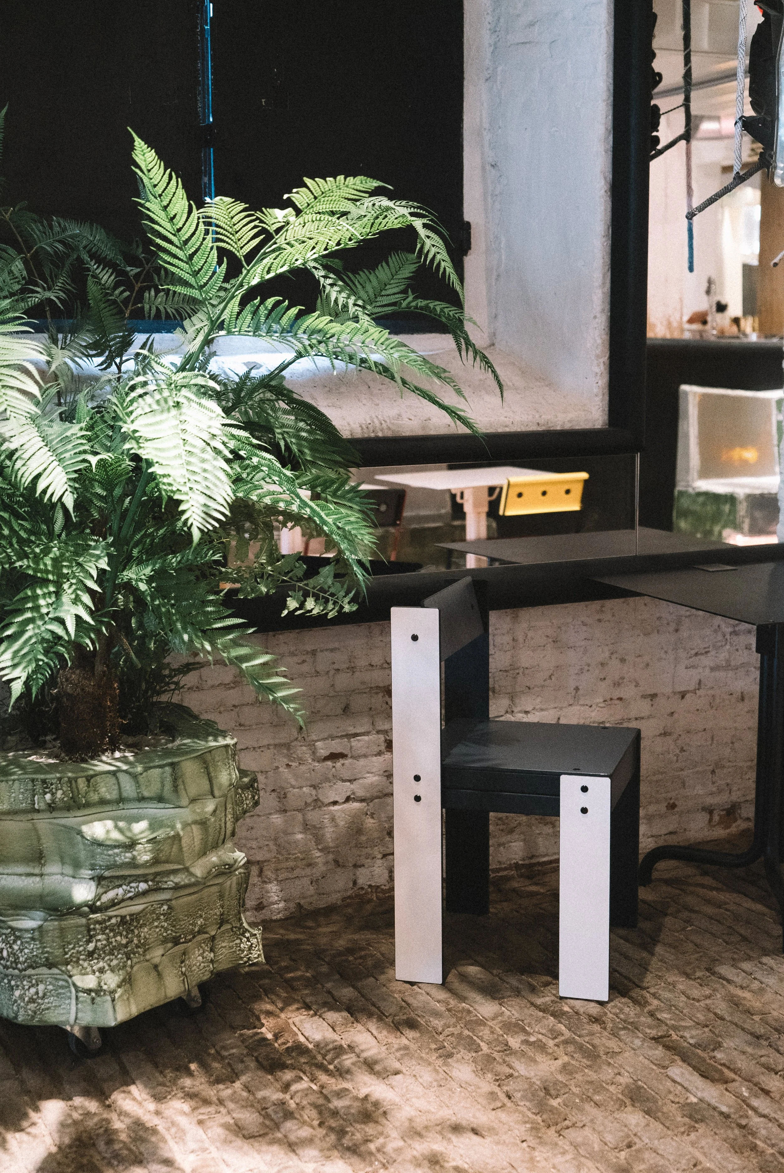 A large green potted fern plant sitting on a marble and metal plant stand next to a black and white modern chair in a room with a brick floor and white brick wall.