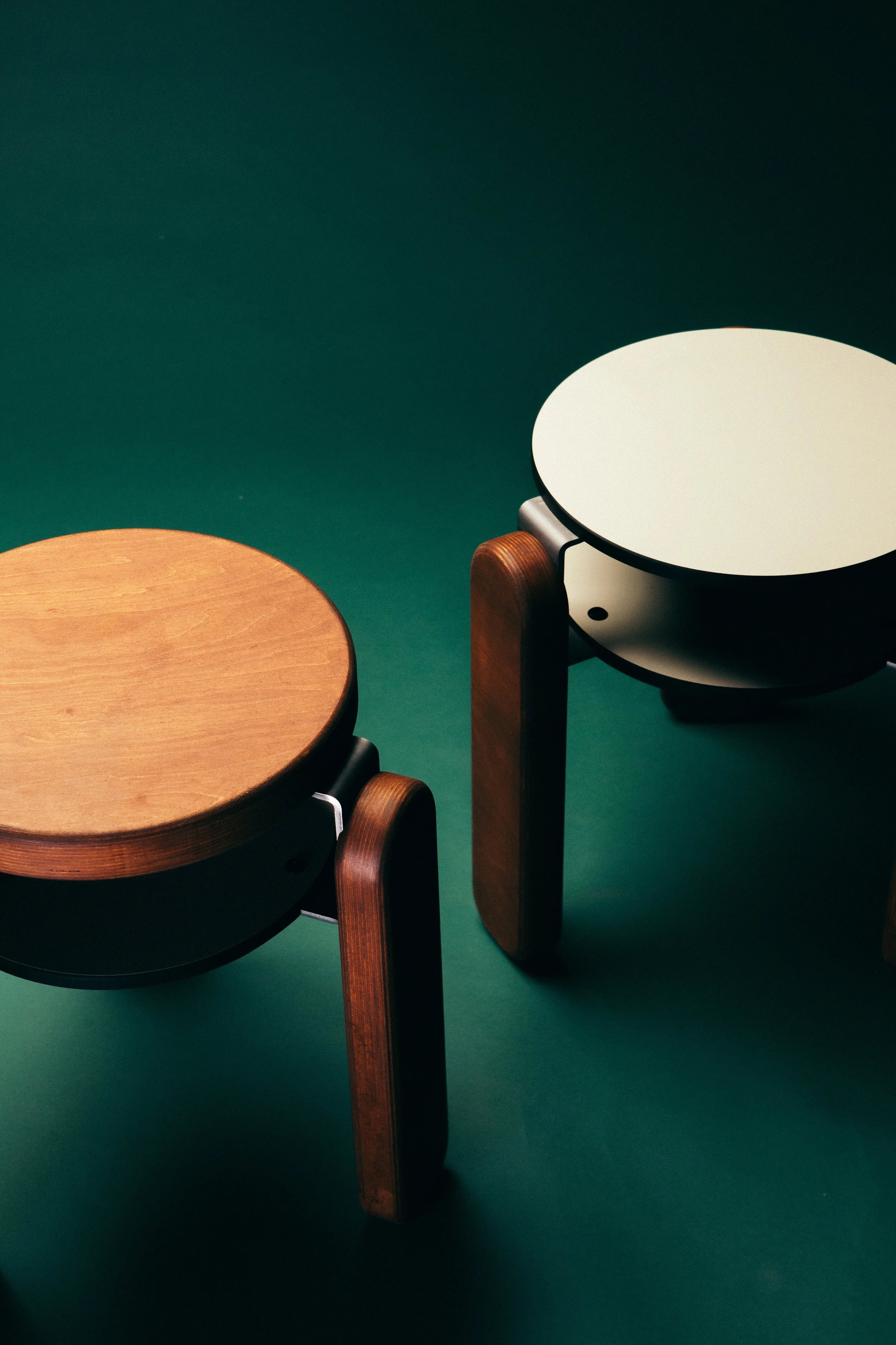 Two modern wooden stools with round tops, one with a natural wood finish and the other with a white top, set on a green background.