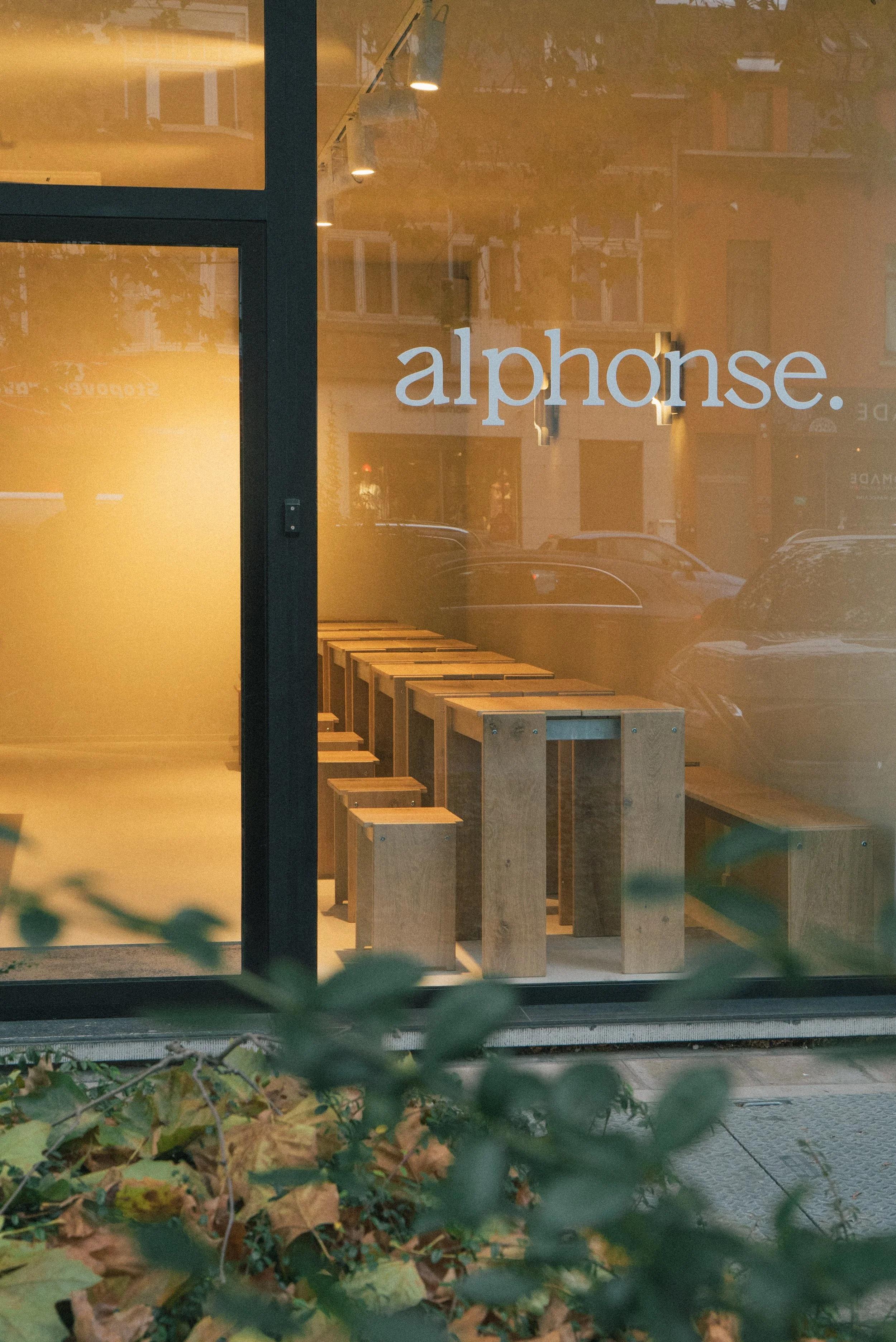 A storefront window with the word 'alphonse.' written on it. Inside, wooden tables and stools are visible. The window reflects a street scene with parked cars and buildings.