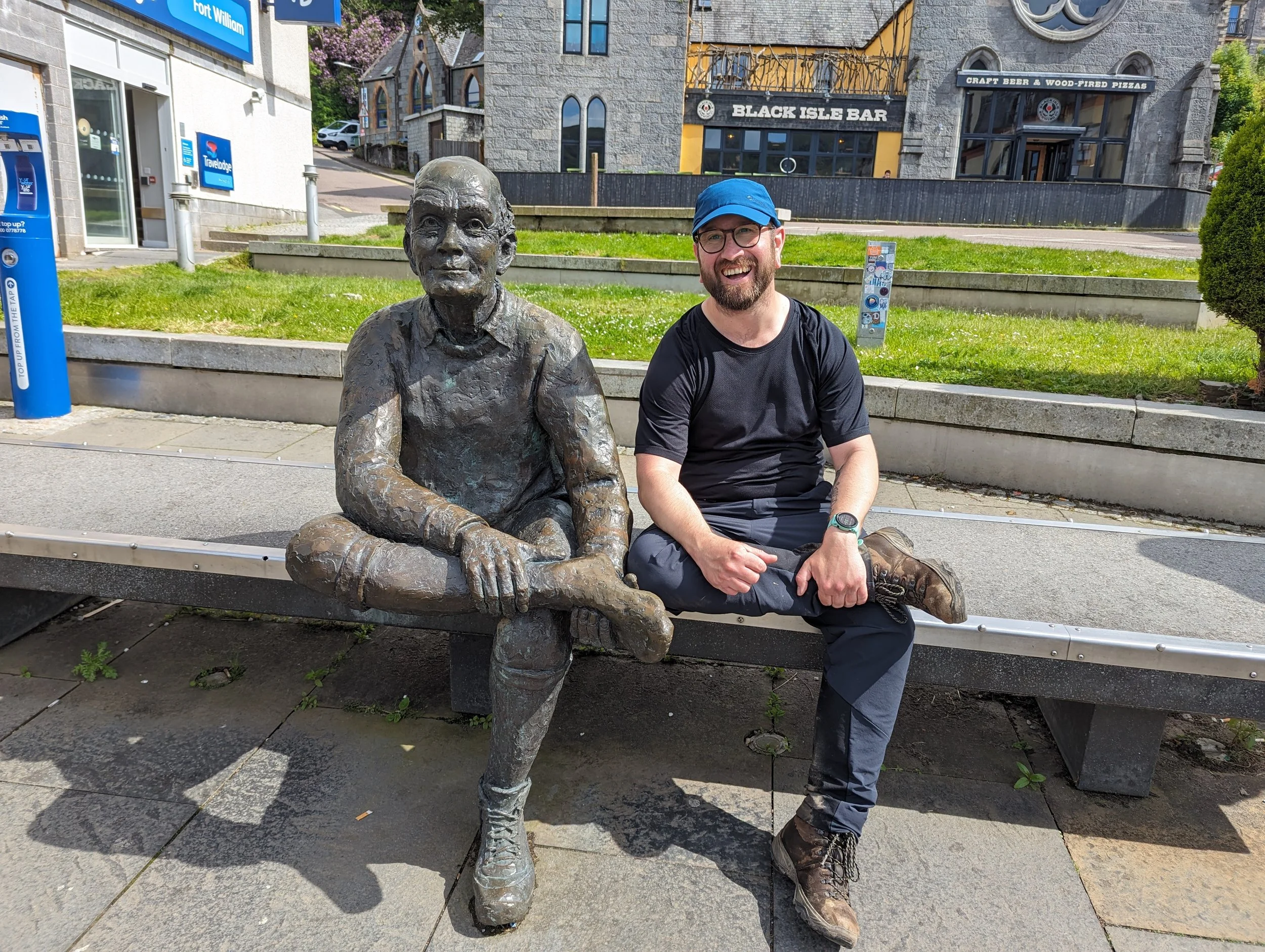 A man with a blue cap and glasses sitting on a park bench next to a bronze statue of a person sitting cross-legged with hands on knees. Buildings and signs in the background including 'Black Isle Bar' and 'Craft Beer & Wood-Fired Pizzas'.