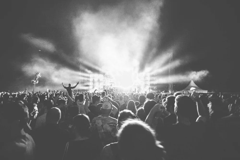 A large crowd of people at a concert or music festival, with stage lights shining brightly in the background. The photo is in black and white.
