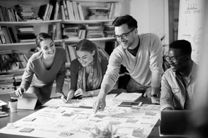 A group of five young people collaborating at a table covered with papers, notebooks, and a laptop in a library or office, all smiling and engaged in discussion.