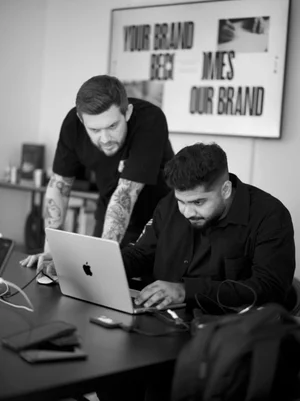 Two men working together at a table with laptops, one sitting and the other standing, in a room with a sign on the wall that says 'YOUR BRAND'.