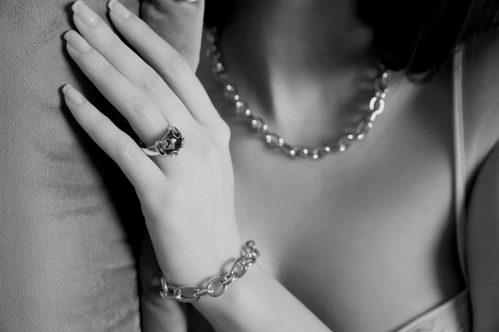 A close-up black and white photo of a woman wearing jewelry, including a chain necklace, a bracelet, and a ring, with her hand resting on her shoulder.