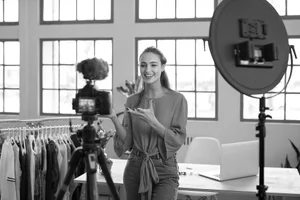 A young woman is filming a video or taking a photo in a bright room with large windows, standing beside a table with a laptop and clothes on a rack, with professional lighting and camera equipment set up.
