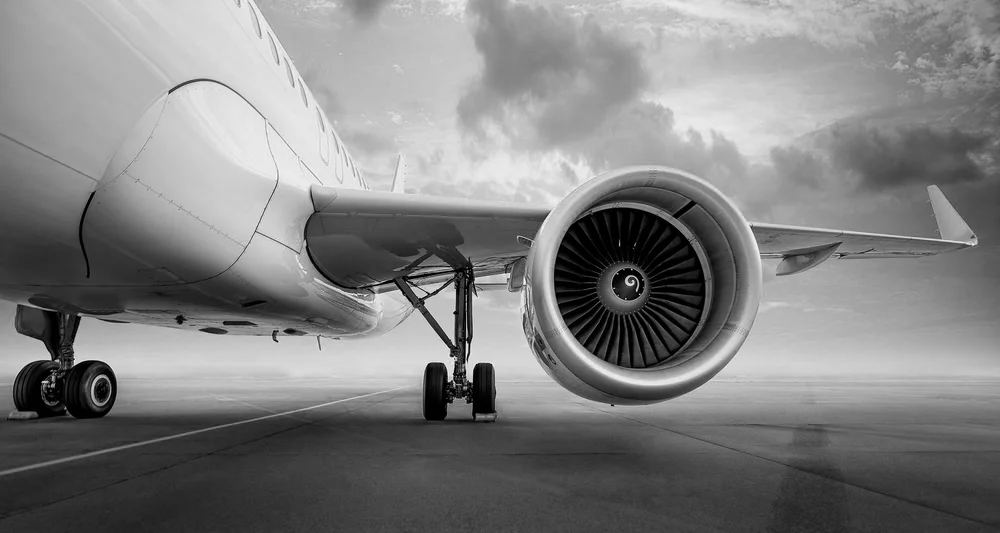 Black and white photo of an airplane on the tarmac, showing the underside of the fuselage, nose, and one engine with the open fan blades visible, aircraft's landing gear extended, and cloudy sky in the background.