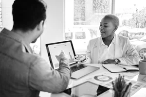 A man and woman having a business discussion in an office, with the woman presenting data on a laptop screen showing bar and pie charts.