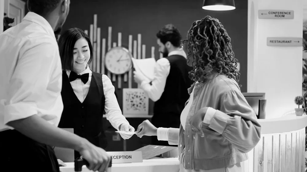 A woman at a reception desk handing a card or paper to a man. Another woman, possibly a staff member, is smiling and looking at the transaction. In the background, a man is reading a book or menu near a clock, with signs pointing to a conference room and restaurant.