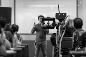 A person giving a presentation in front of an audience in a classroom or seminar room, with a camera recording the event.