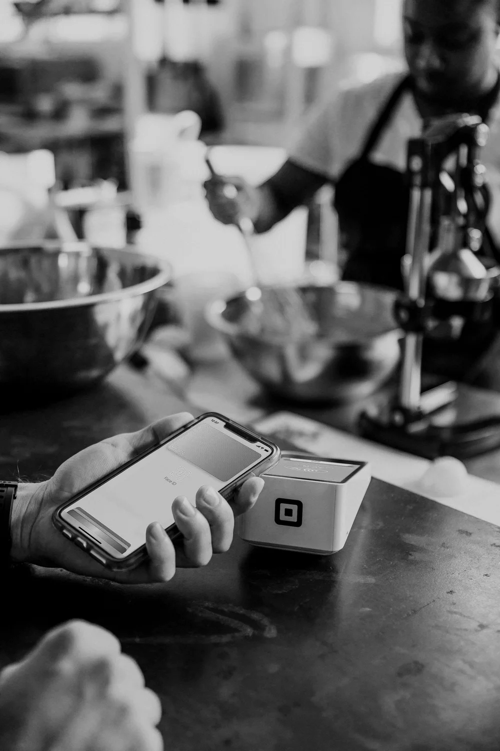 A person holding a smartphone with a QR code scanner app open, scanning a Square payment device on a wooden table in a kitchen or bakery setting.