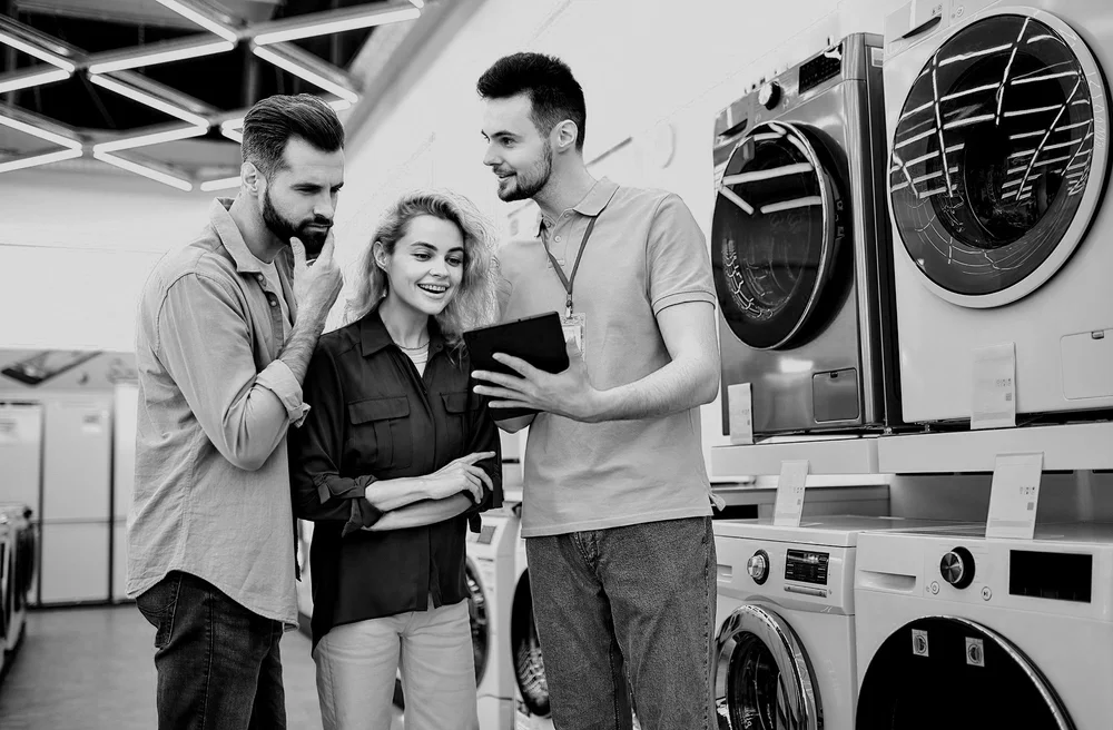Three people, two men and one woman, looking at a tablet in an appliance store, surrounded by washing machines and dryers.