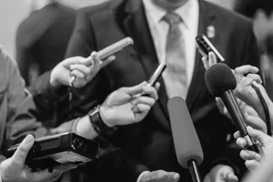 A person in a suit is being interviewed, surrounded by reporters holding microphones and recording devices.