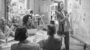 A woman in business attire presents a flowchart on a glass whiteboard to a group of four colleagues, some seated at a table and others standing, in an office with large windows and bookshelves.