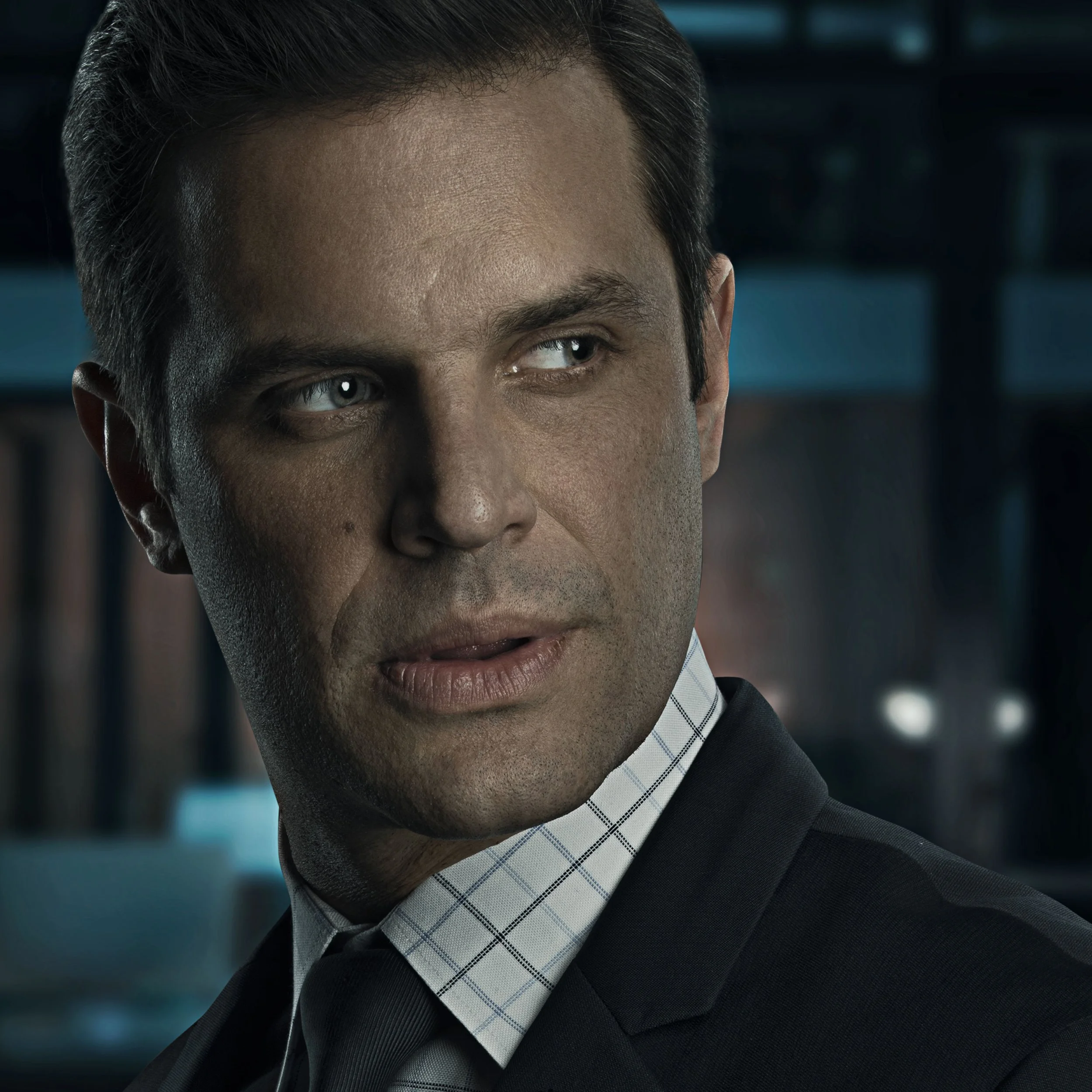 Close-up of a man with slicked-back hair, wearing a dark suit, white dress shirt with a checkered pattern, and black tie, looking to his right with a serious expression.