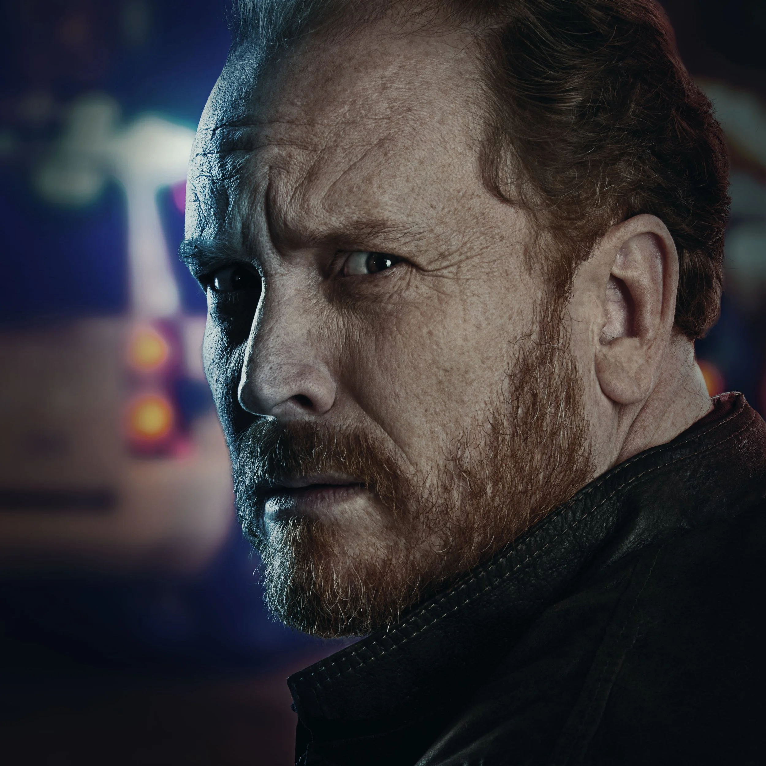 Close-up of a man with reddish hair and beard, looking over his shoulder with a serious expression, with blurred colorful neon or city lights in the background.
