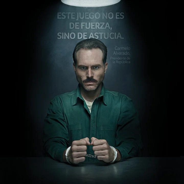 Man with serious expression, wearing green jacket, holding handcuffs, in front of background with Spanish text and a spotlight.
