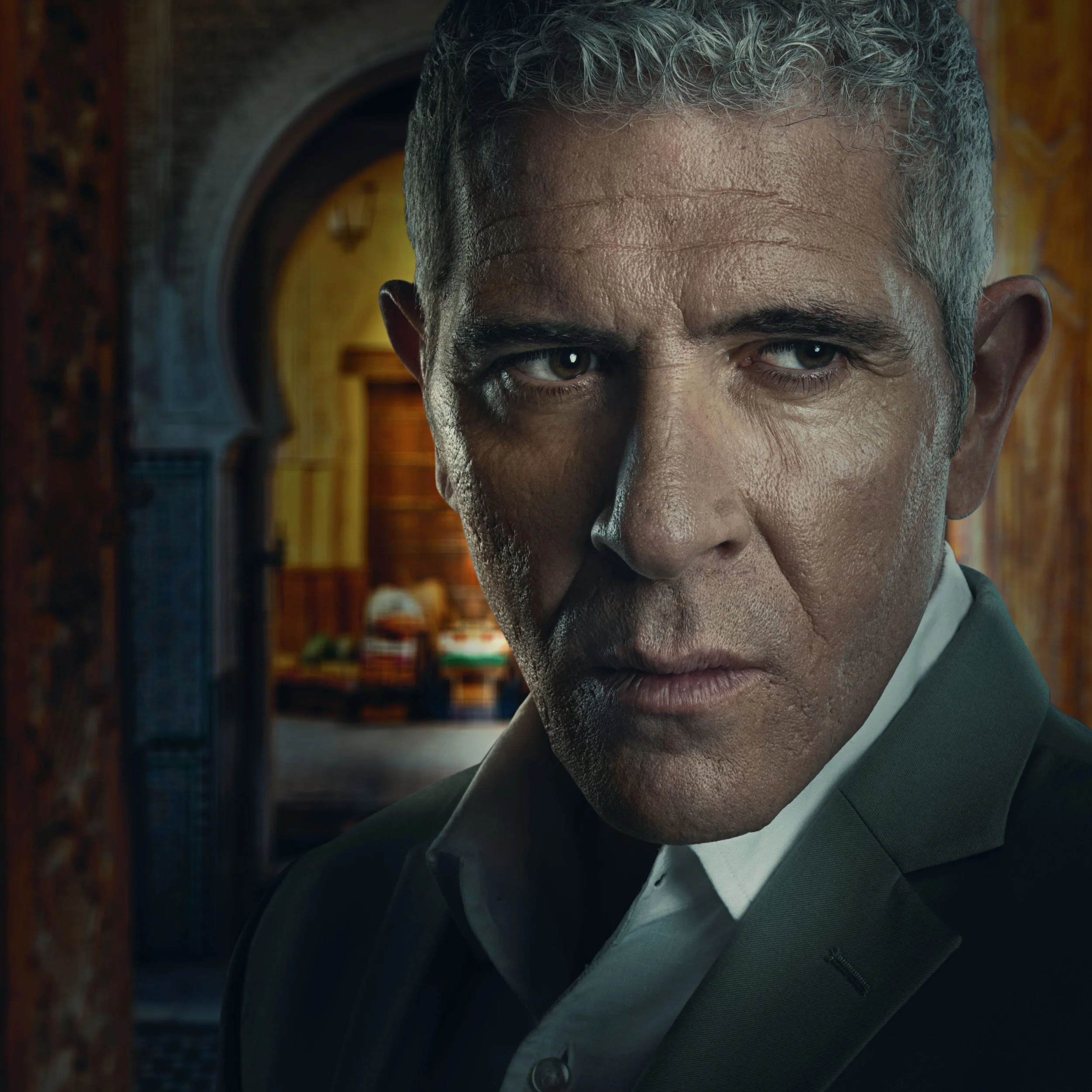Close-up of a middle-aged man with gray hair and piercing blue eyes, wearing a dark suit jacket, inside a richly decorated room with warm wooden paneling and an arched doorway in the background.