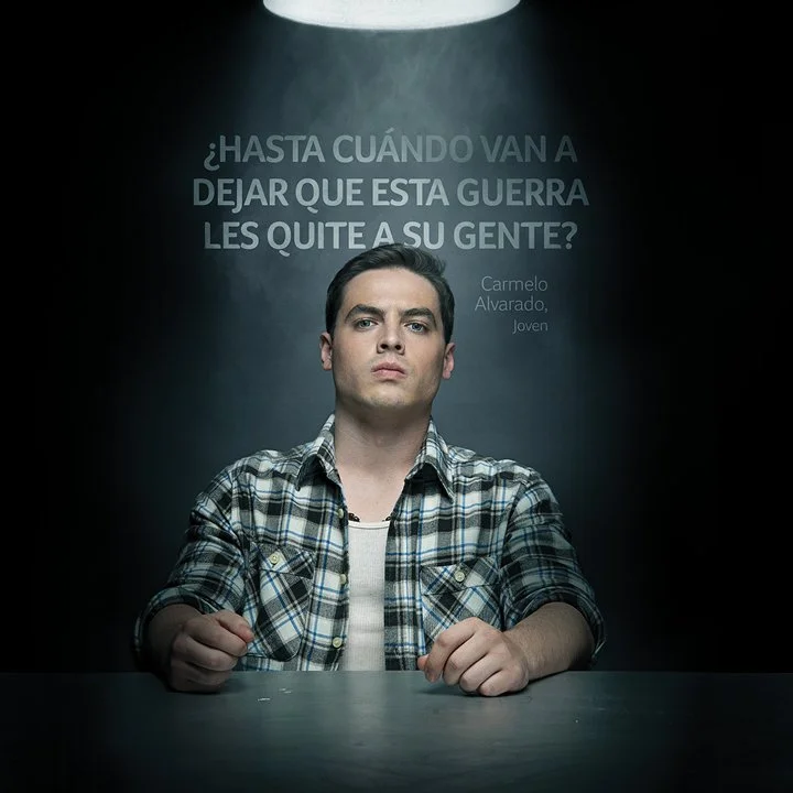 Young man with a serious expression sitting at a table under a spotlight, with a dark background featuring Spanish text questioning how long a war will last, in a documentary or interview style setting.