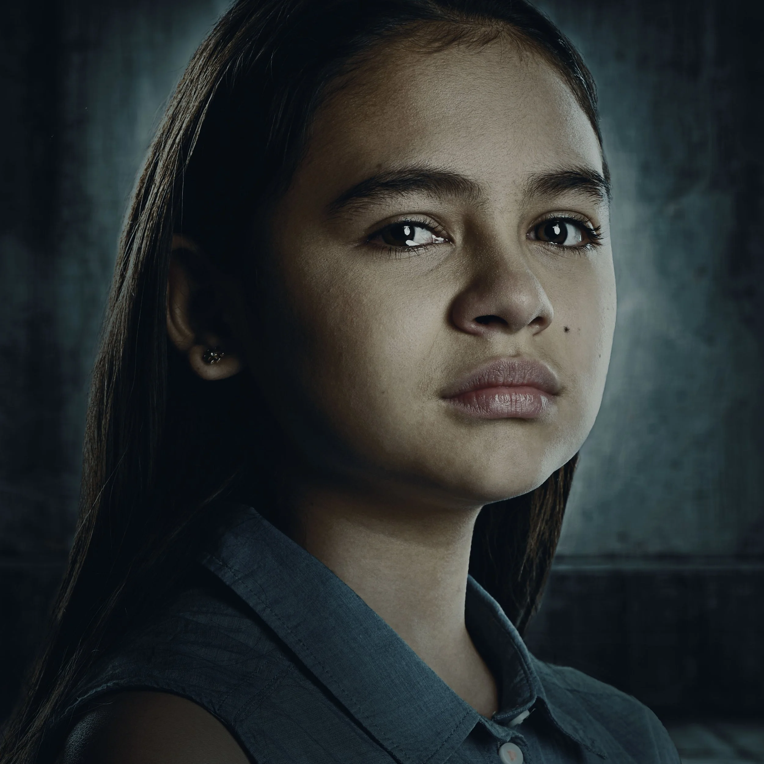 Close-up portrait of a young woman with straight brown hair, wearing a dark shirt, against a dark background.