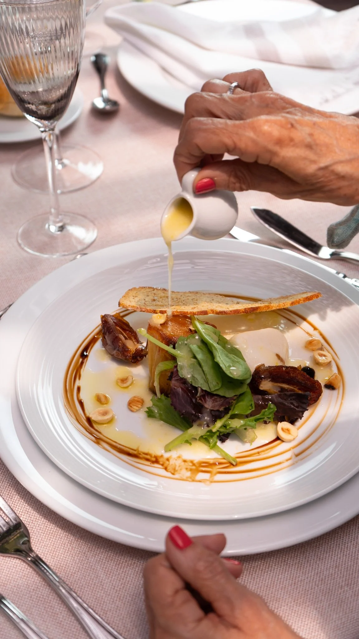Persona rociando salsa sobre un plato con ensalada, ingredientes y pan tostado en un ambiente de restaurante elegante.