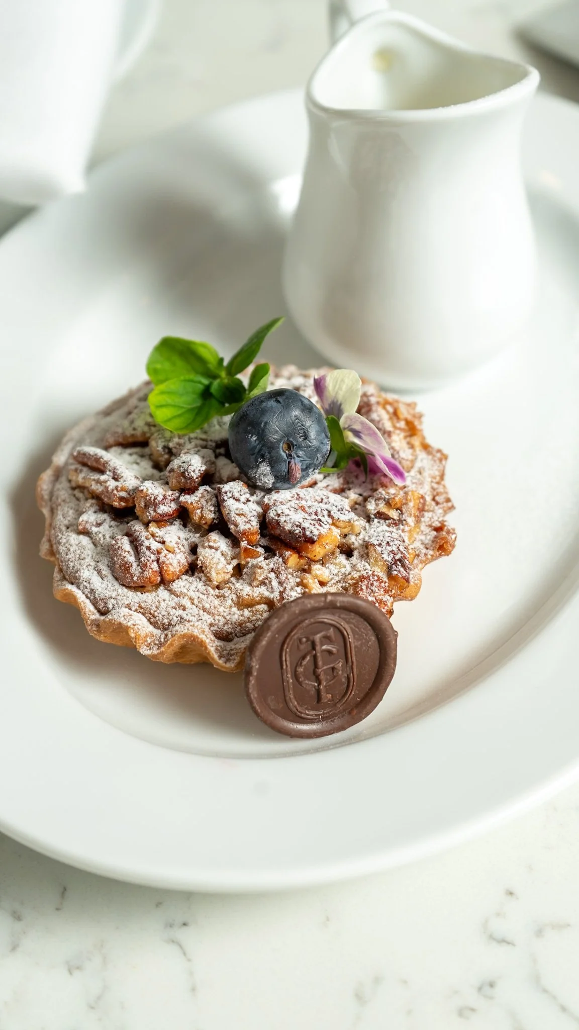 Pastel de nueces con polvo de azúcar, decorado con una mora, una hoja de menta y una flor, servido en un plato con una pequeña jarra de leche.