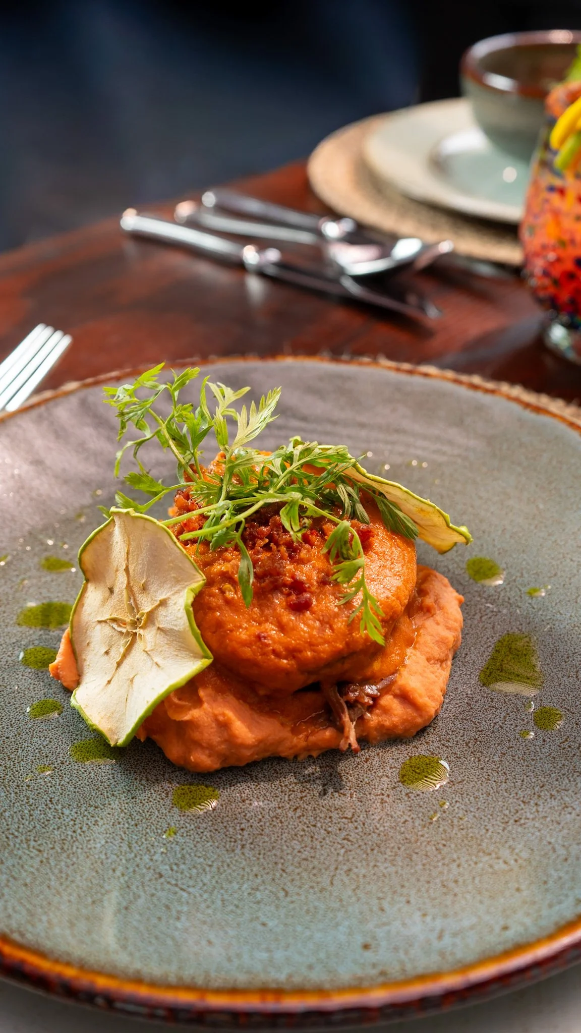 Plato de comida con una albóndiga decorada con hierbas, acompañada de rodajas de fruta y una salsa, sobre un plato de cerámica en un ambiente de comedor.