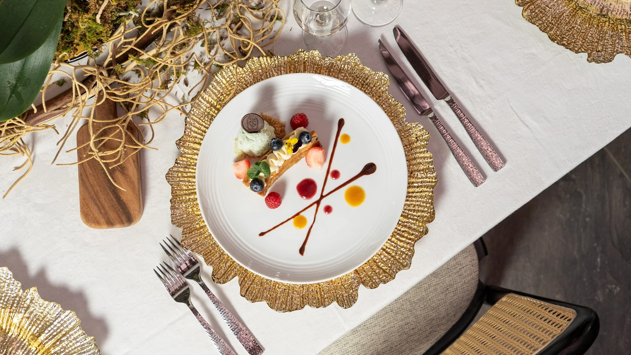Postre decorado con frutas y salsa, servido en un plato blanco con borde dorado, en una mesa con mantel blanco y cubiertos de color rosa, en un ambiente elegante.