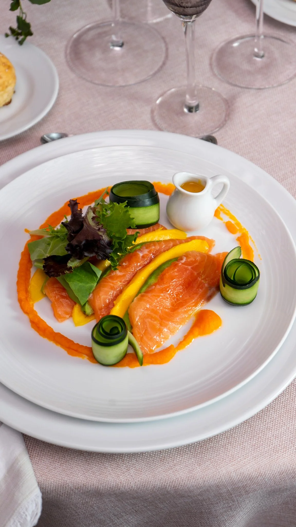 Plato con salmón, ensalada, rodajas de pepino enrolladas y salsas, en un plato blanco sobre una mesa con copas de vino.