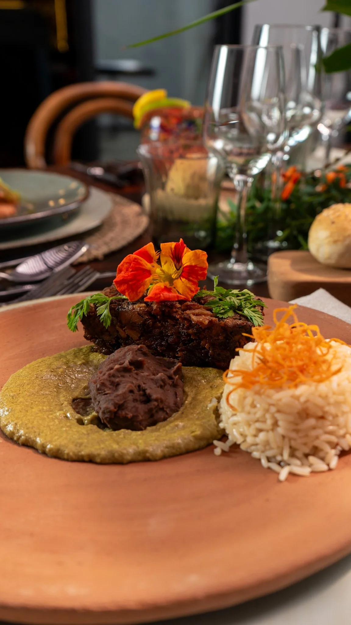 Plato de comida con pollo, arroz, salseada y decorado con flor comestible y perejil, en una mesa con vasos y candelabros en el fondo.