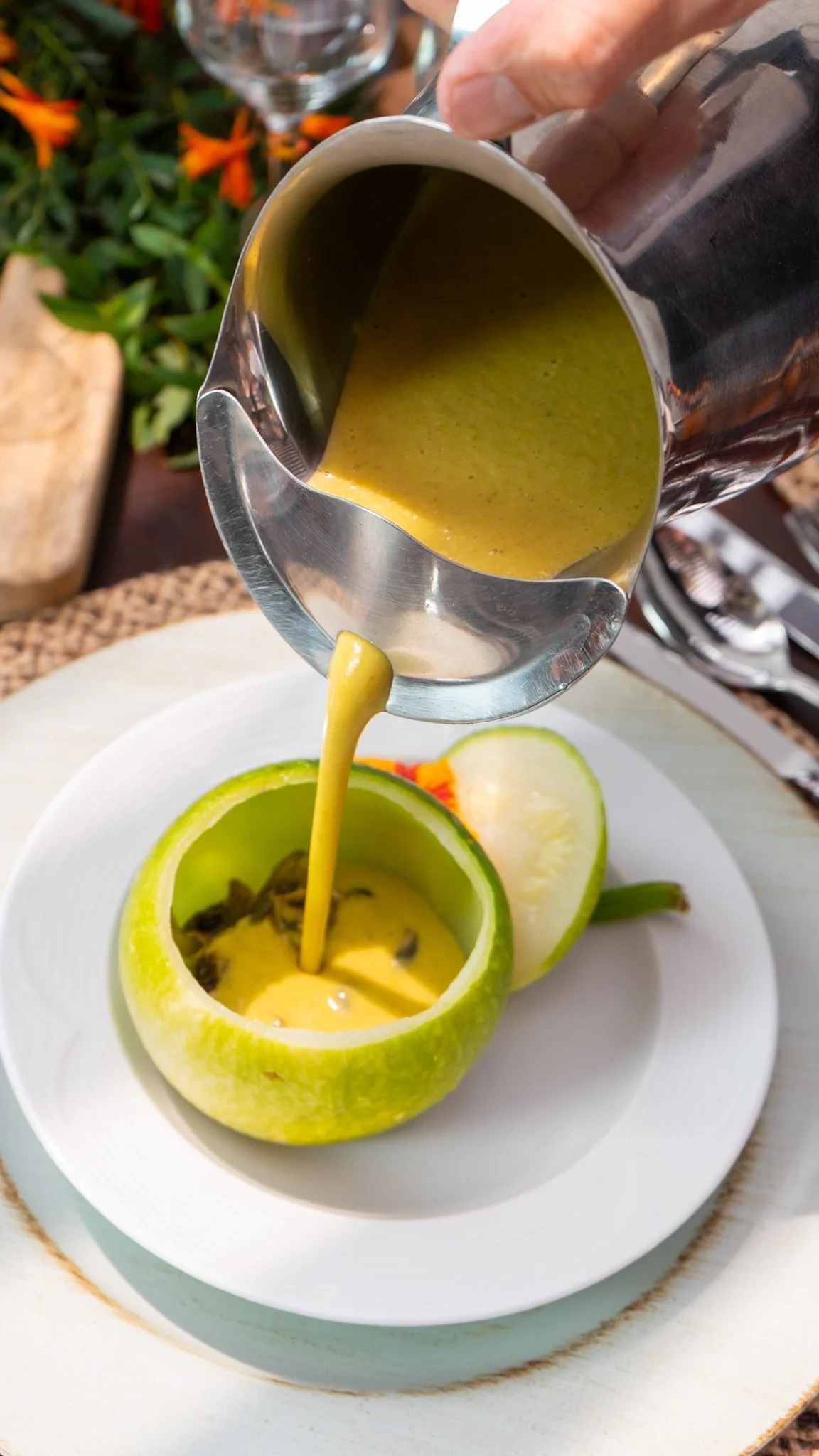 Cuchara vertiendo salsa de aguacate en un plato con un aguacate cortado por la mitad y verduras decorativas.