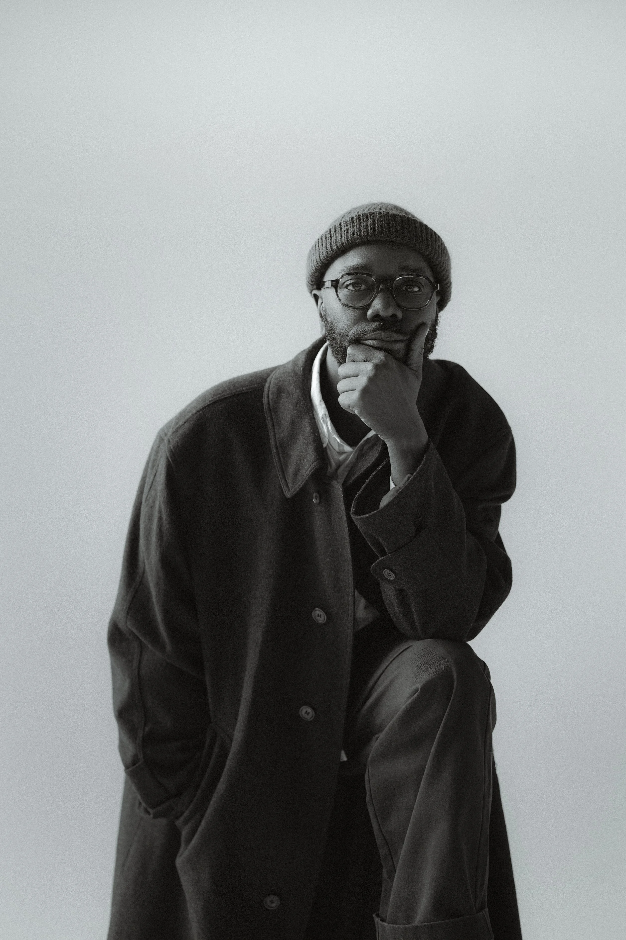 A black-and-white photo of a man wearing glasses, a beanie, a dark coat, and pants, in a contemplative pose with his hand on his chin, sitting against a plain background.