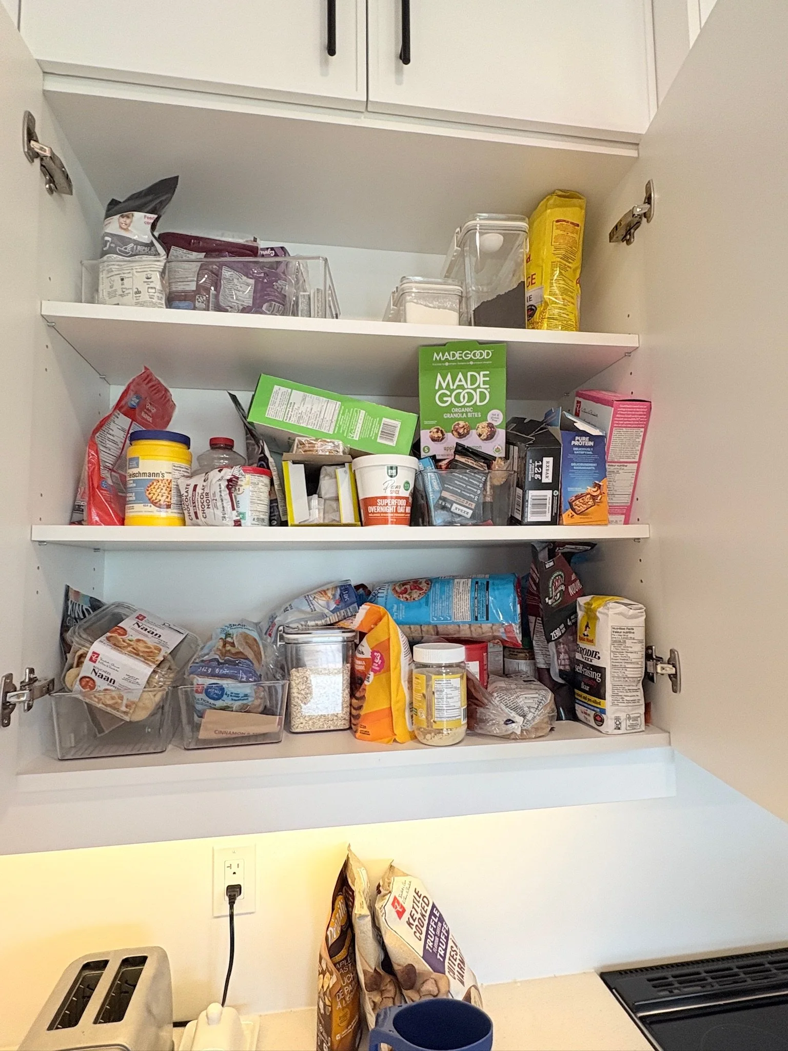 Open kitchen cabinets with various food items, pantry supplies, and kitchen appliances on the counter, including a toaster and a coffee maker.