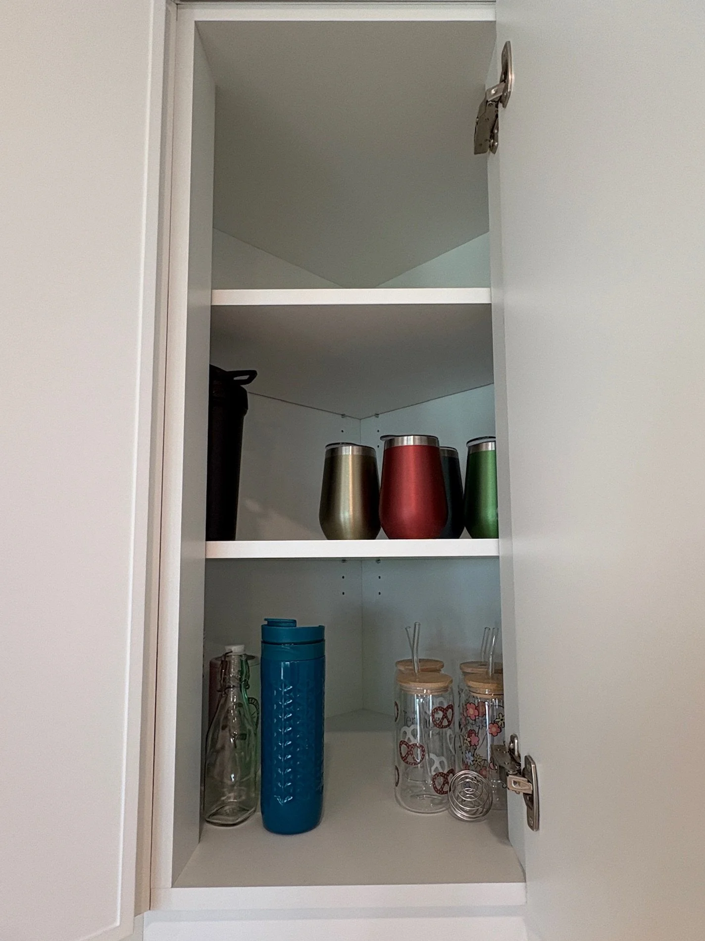 Open kitchen cabinet with various cups, glasses, and bottles inside.