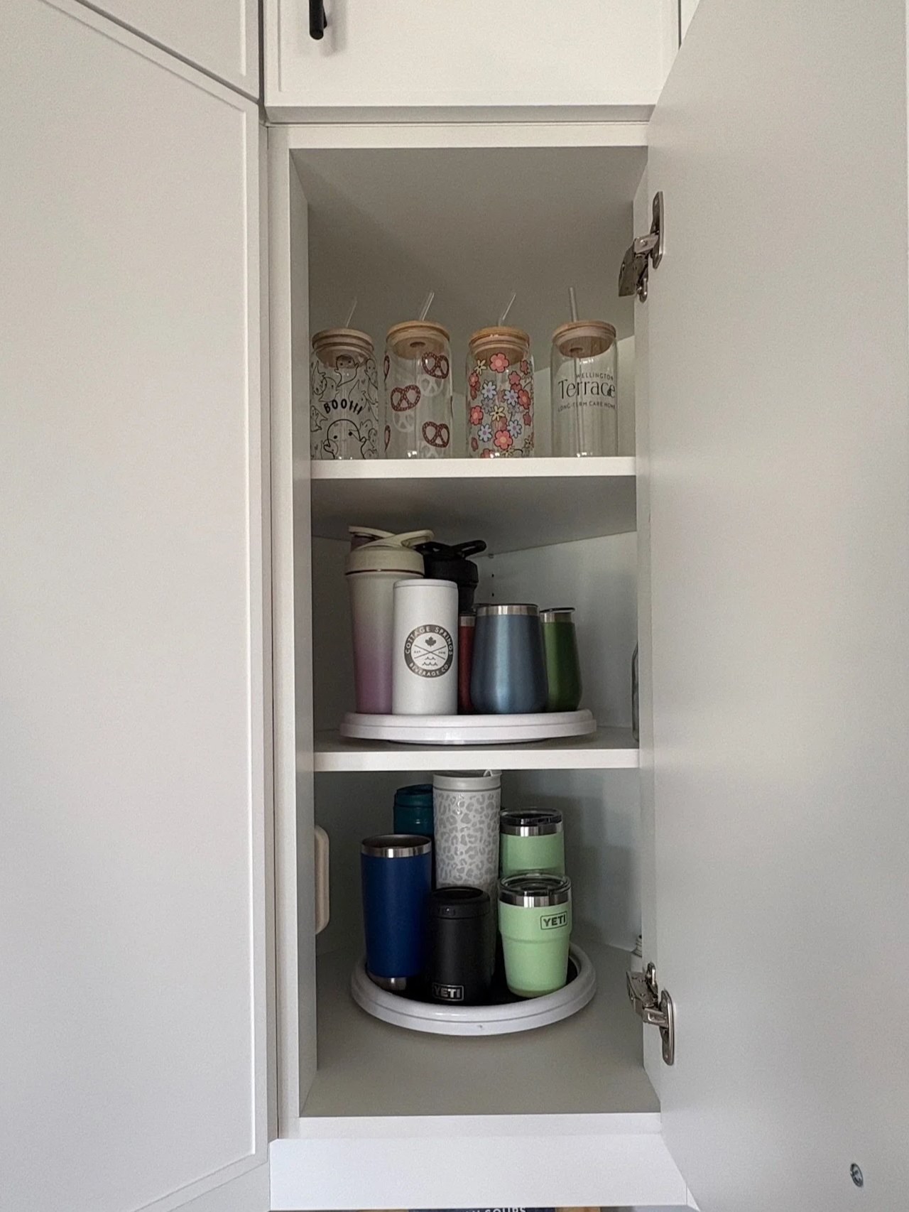 Open kitchen cabinet with three shelves filled with various travel mugs and tumblers of different colors and designs.