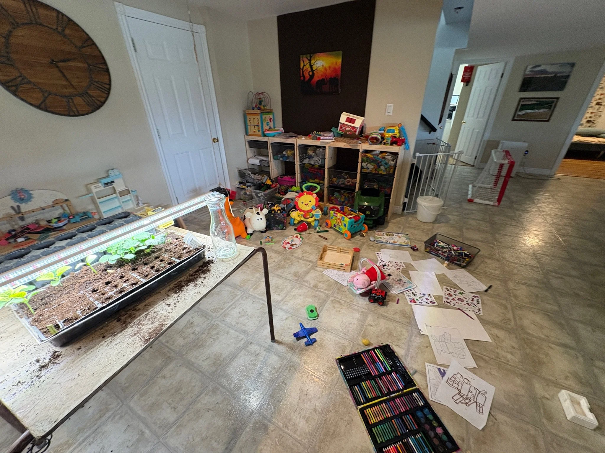 A cluttered room with various toys, papers, and art supplies scattered across the floor. There is a fish tank on a table with plants growing inside. A large wall clock is on the wall, and a painting of elephants at sunset hangs on one wall. A white b