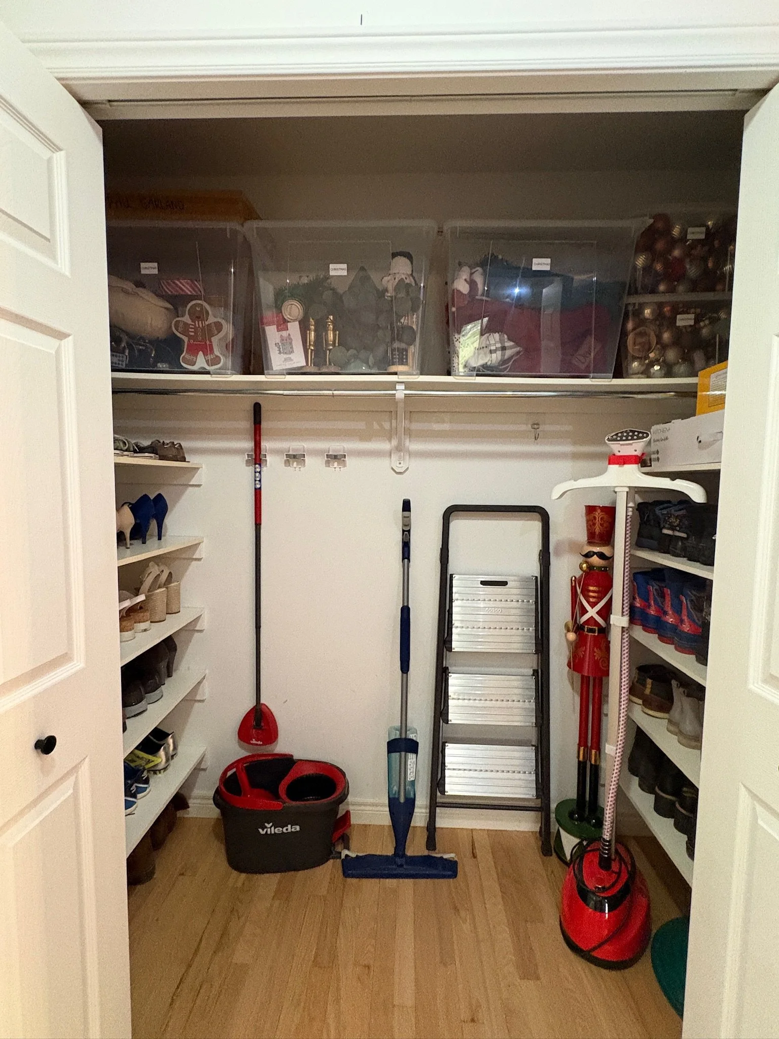 A closet with shelves holding shoes and storage bins, and cleaning supplies including a vacuum, broom, and dustpan.