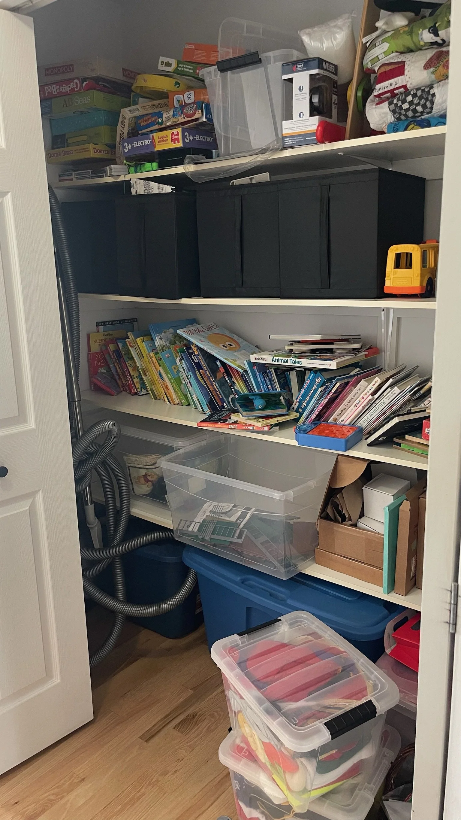 Closet filled with shelves of books, magazines, plastic storage bins, board games, and miscellaneous household items, including a toy school bus and a vacuum hose.