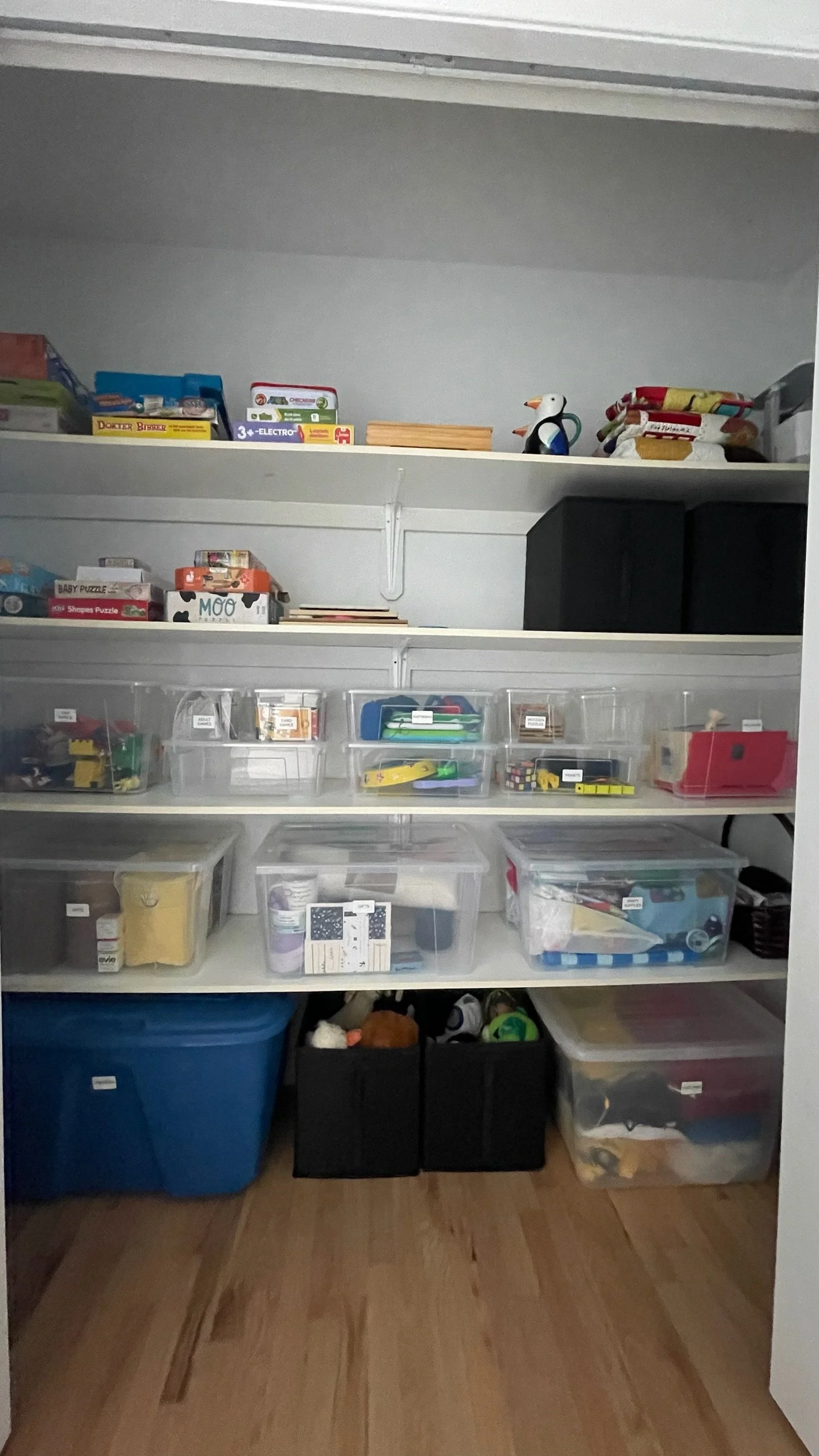 A closet with white shelves filled with plastic storage bins, boxes, and toys. Top shelf has board games and a penguin figurine. Lower shelves hold organized bins with toys and miscellaneous items. The floor is wooden.