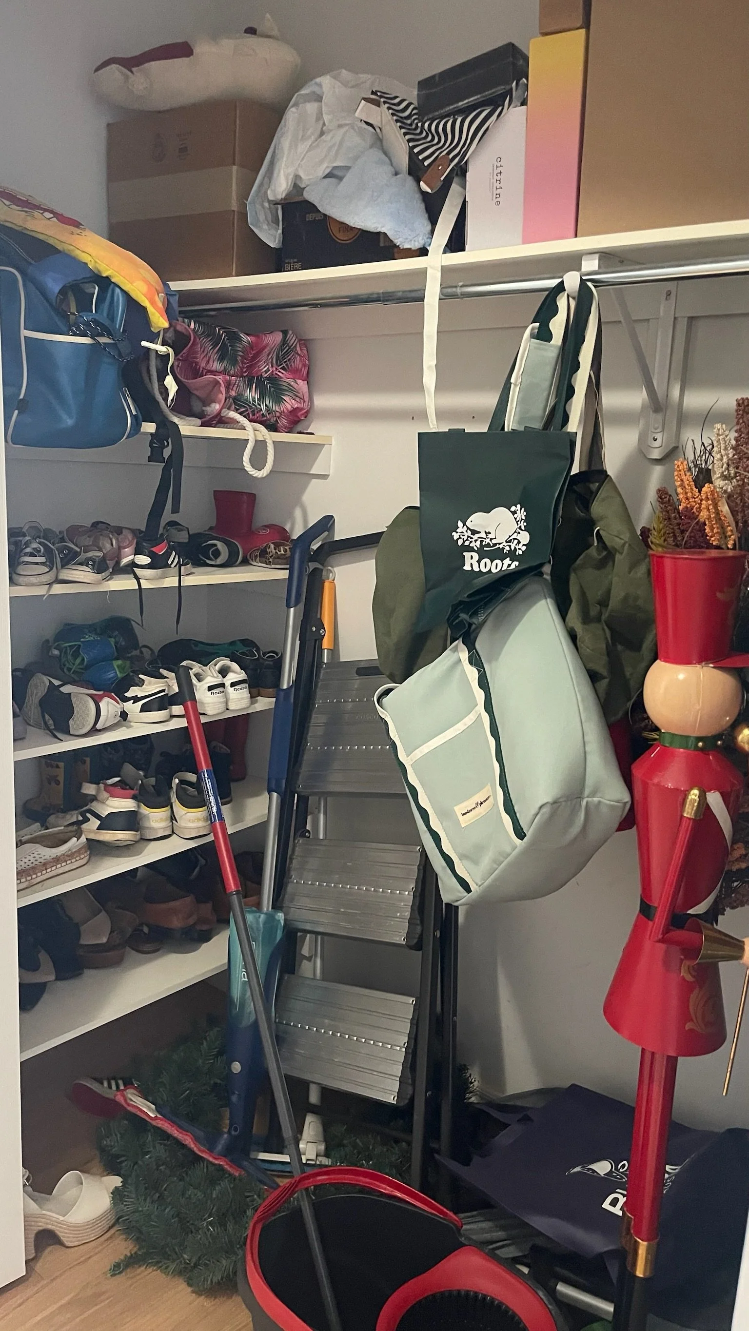 A cluttered closet or storage area with shoes, boxes, bags, and decorations including a small evergreen tree. There are also cleaning tools, a step ladder, and a decorated red and gold item resembling a Christmas stocking holder.