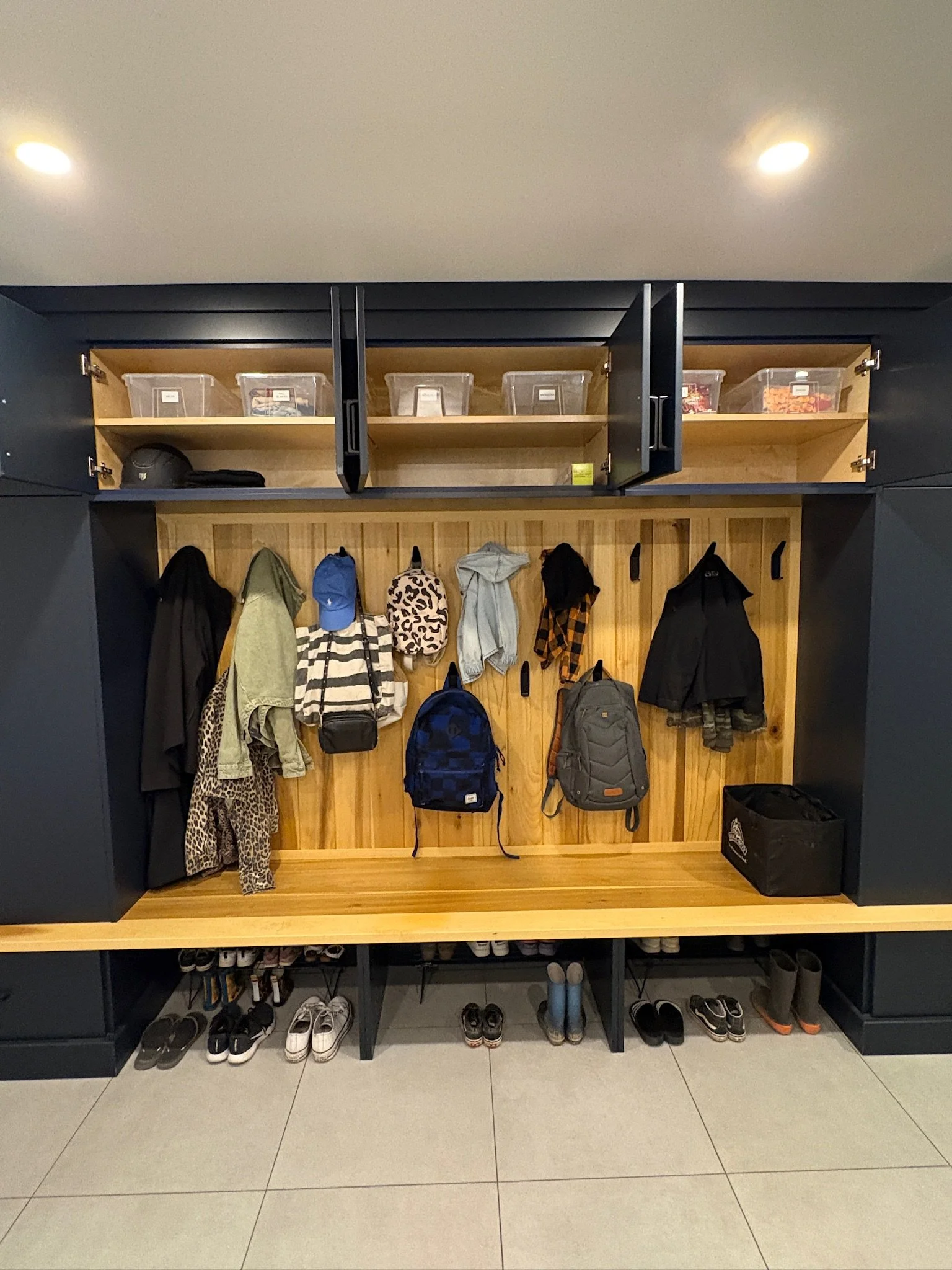 A hallway coat and shoe storage area with hooks, hooks with hanging jackets and backpacks, shelves with hats and boxes, shoes on the bottom shelf, and a black storage bin on the floor.